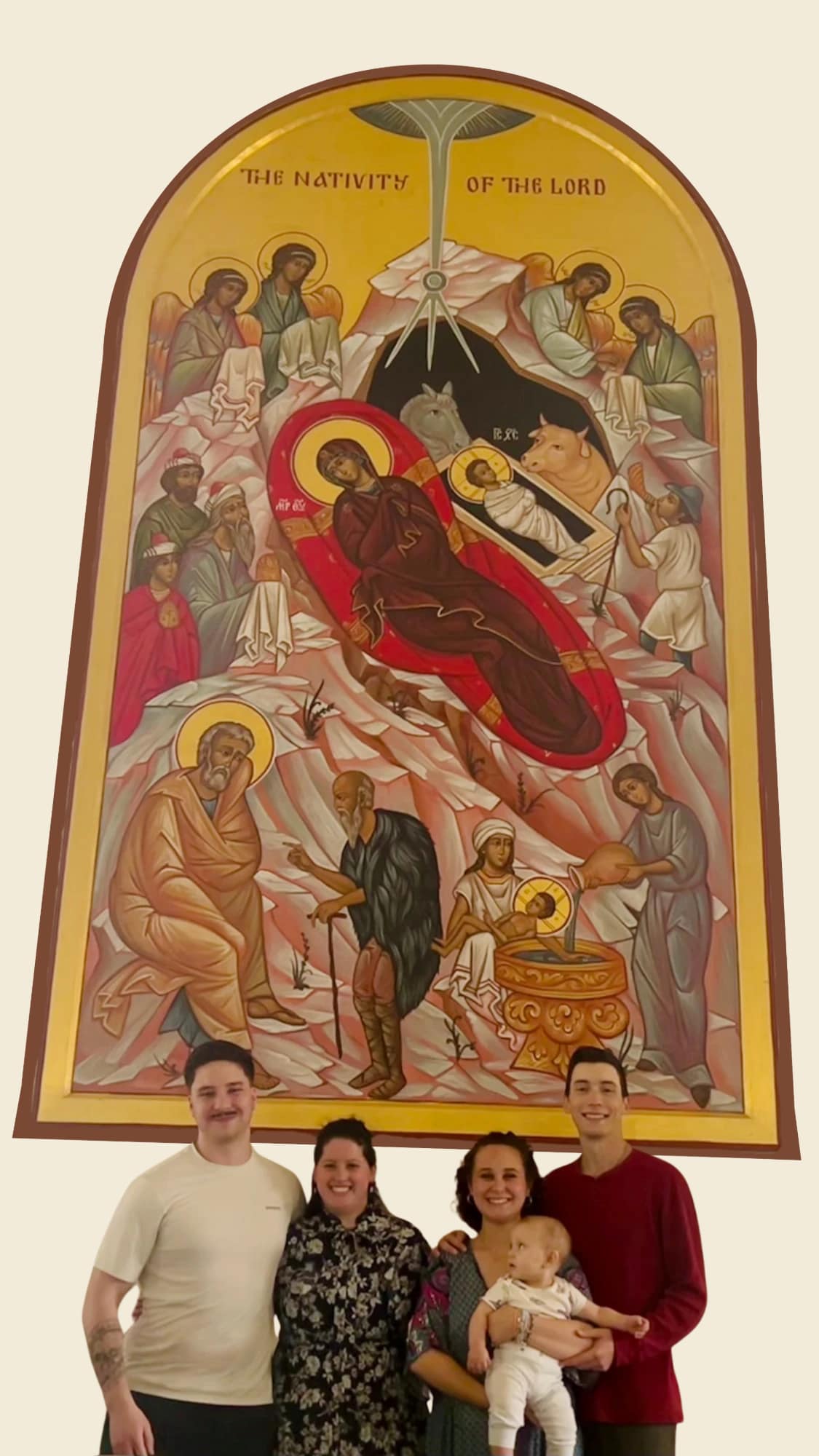 Four smiling adults — one of them holding an infant — stand together beneath a tall, gold-grounded icon of the Nativity of the Lord, painted with angels, the Magi, the Mother of God and the Christ Child in the manger.